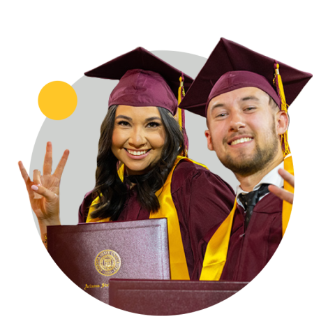 Two ASU grads where maroon and gold graduation regalia and hold "forks up."
